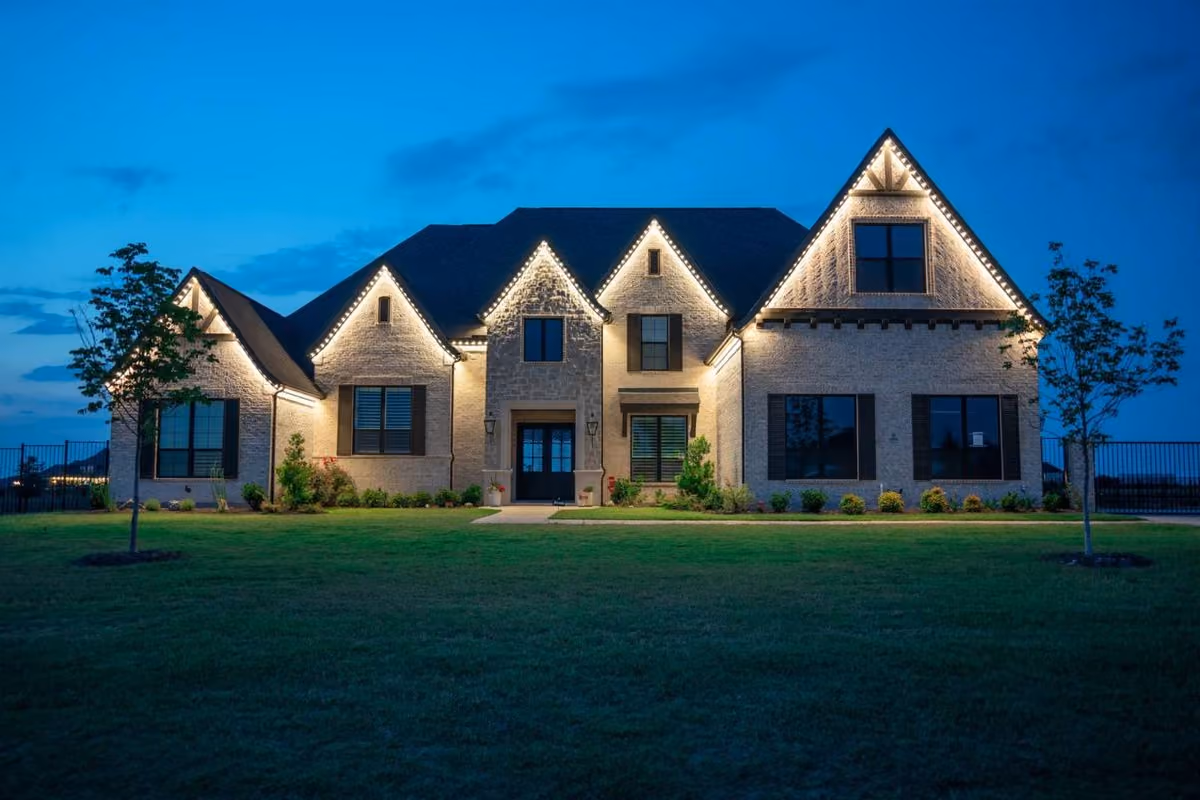 Large modern house at dusk with beige brick exterior and peaked rooflines outlined by warm white accent lighting, surrounded by green lawn and small trees.