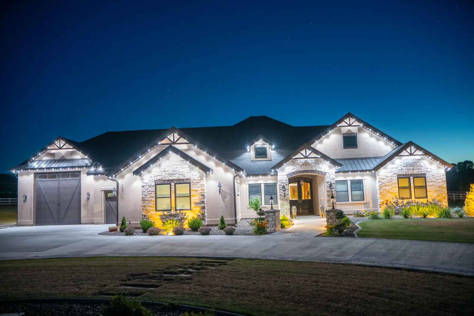 Large modern house at night lit with white string lights along the roof edges and warm yellow ground lighting highlighting stone accents and landscaping, with a wide driveway and neatly trimmed lawn in front.