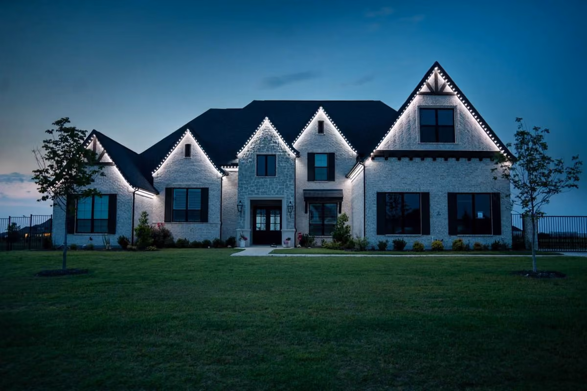 Large white brick house at dusk with peaked rooflines decorated with white string lights along the edges, black-trimmed windows, a well-maintained lawn, and two small trees on each side.