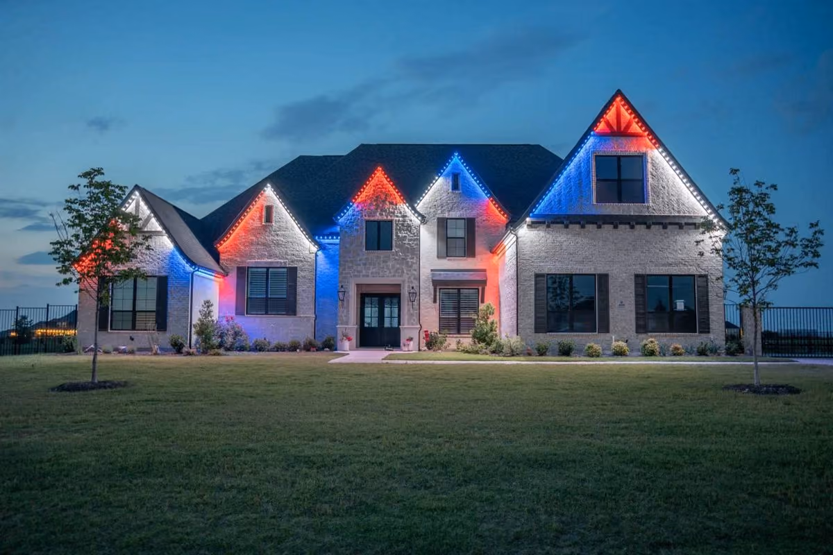 Large white brick house at dusk with a dark roof and red, white, and blue holiday lights outlining the roof peaks, surrounded by a green lawn and two small trees.