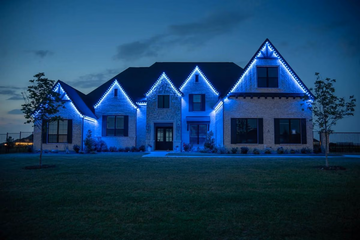 Large modern house at dusk with blue decorative lights outlining the roof edges and multiple gables, front lawn, and two small trees.