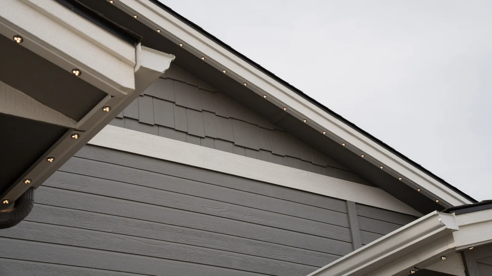 Close-up of a house roof corner featuring gray horizontal and shingle siding with white trim and small round outdoor lights along the eaves.