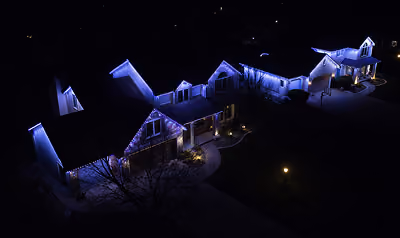 Aerial night view of a row of houses decorated with blue and white Christmas lights outlining the roofs and eaves, with some warm lights near the entrances and a lit street lamp on the sidewalk.