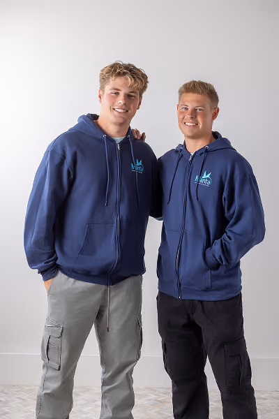 Two young men standing side by side against a plain white background, both wearing navy blue hooded zip-up sweatshirts with a light blue logo on the chest, one with light gray cargo pants and the other with black pants, smiling at the camera.