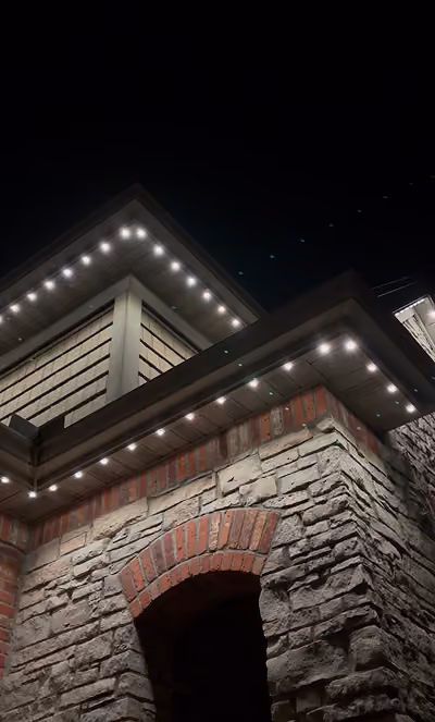 Night view of a stone and brick house corner with white exterior lights lining the roof edges.