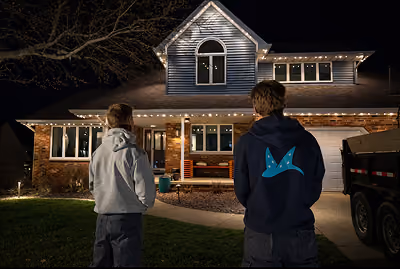 Two people standing outside at night looking at a two-story house decorated with white string lights along the roof edges.