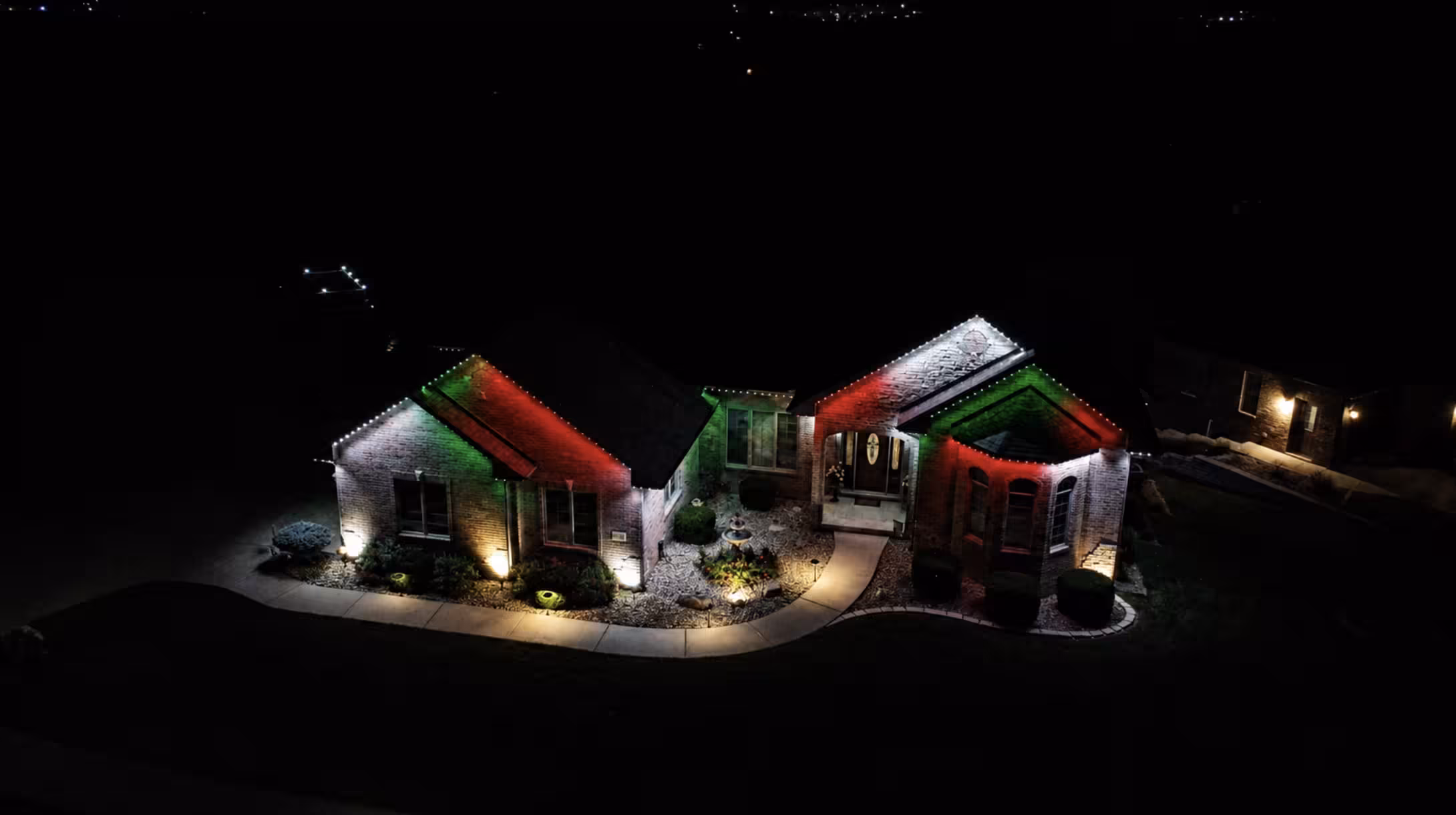 Aerial night view of a house decorated with roof lights in red, green, and white colors and ground spotlights illuminating the front yard and walkways.