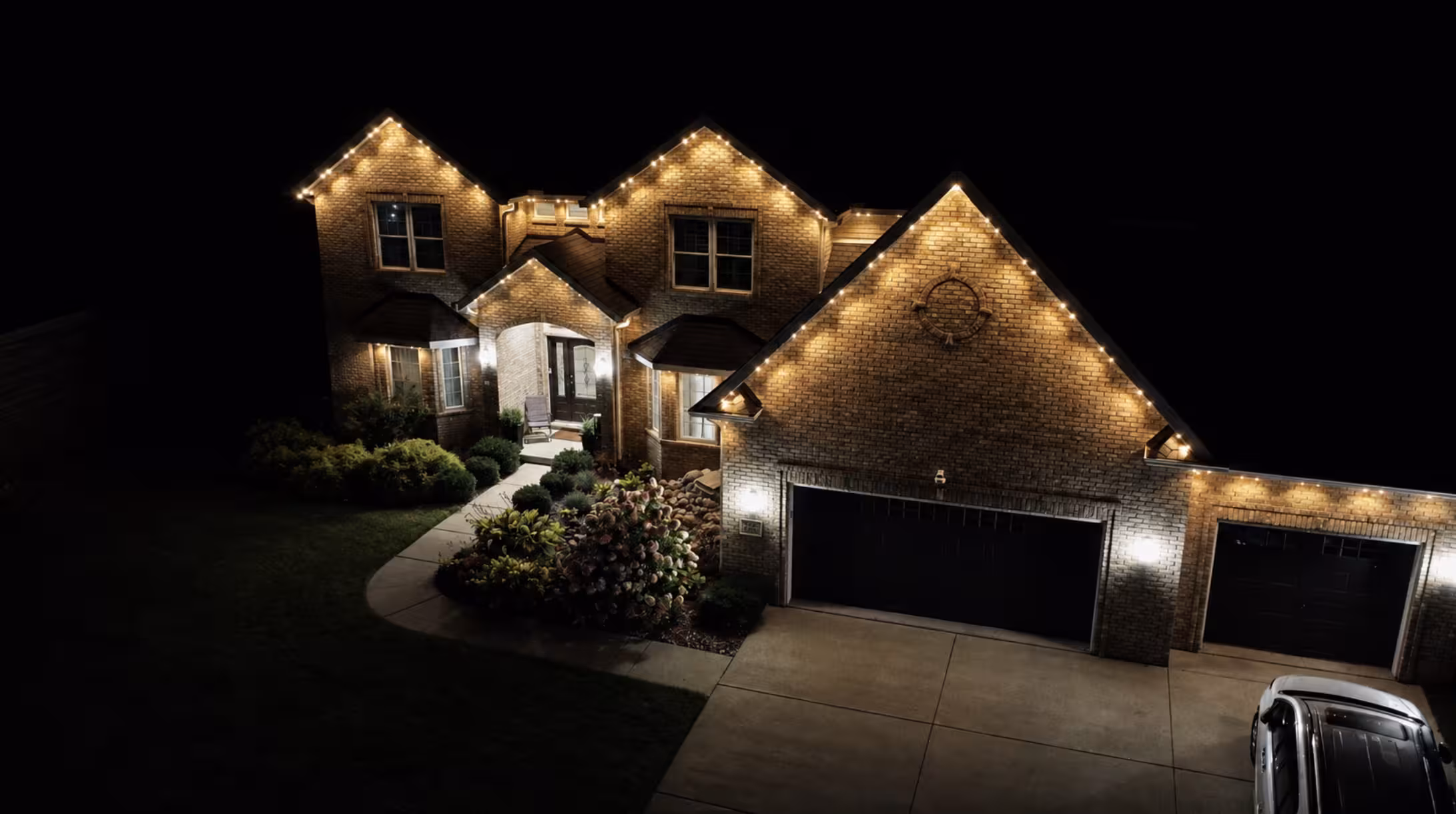 Large brick house at night with illuminated roofline lights outlining the gables and garage, a well-lit entrance, and a driveway with a parked car.