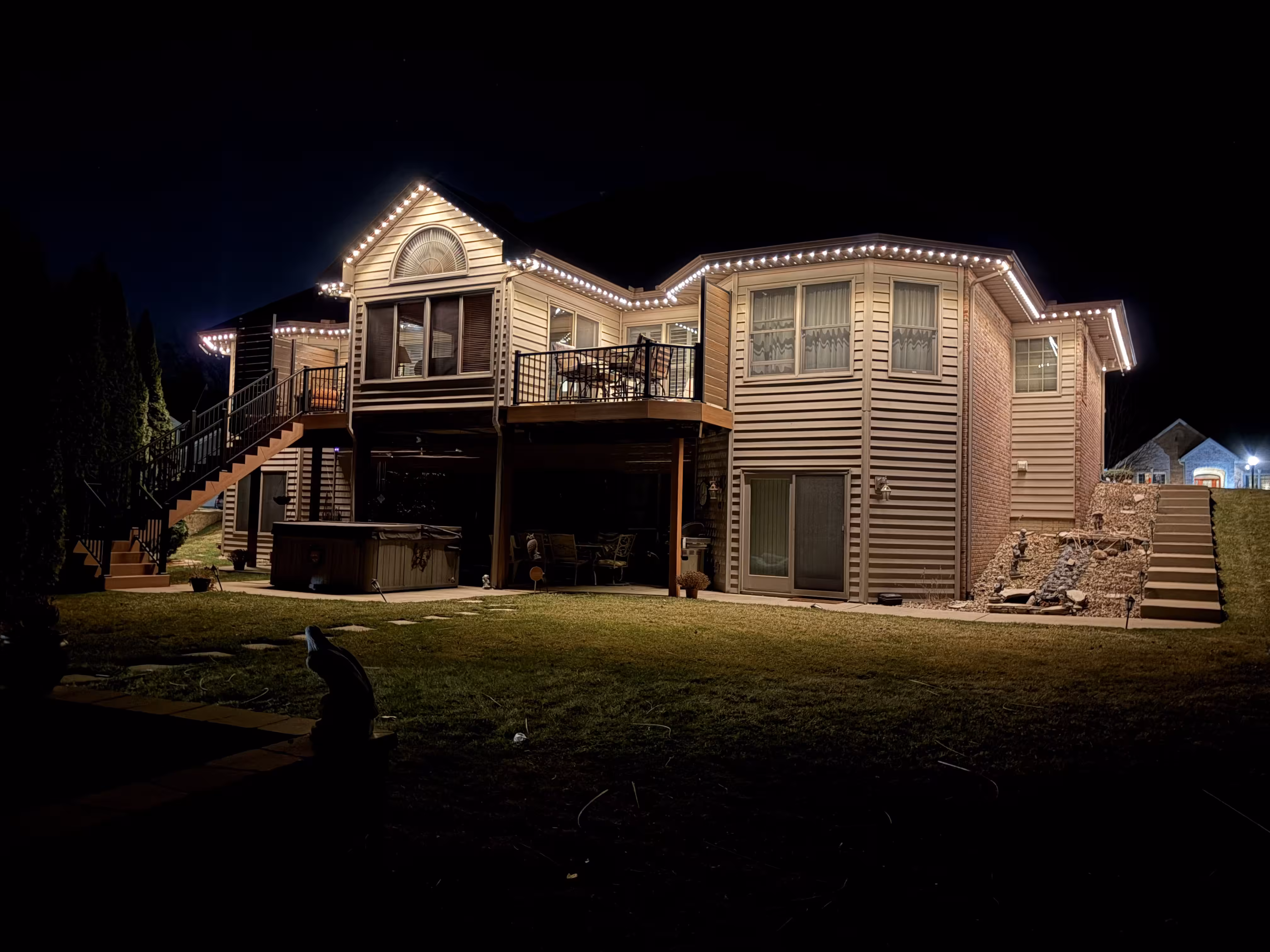 Night view of a two-story house with beige siding and brick accent, decorated with white string lights along the roofline, featuring an outdoor staircase, upper deck with patio furniture, and a grassy backyard.