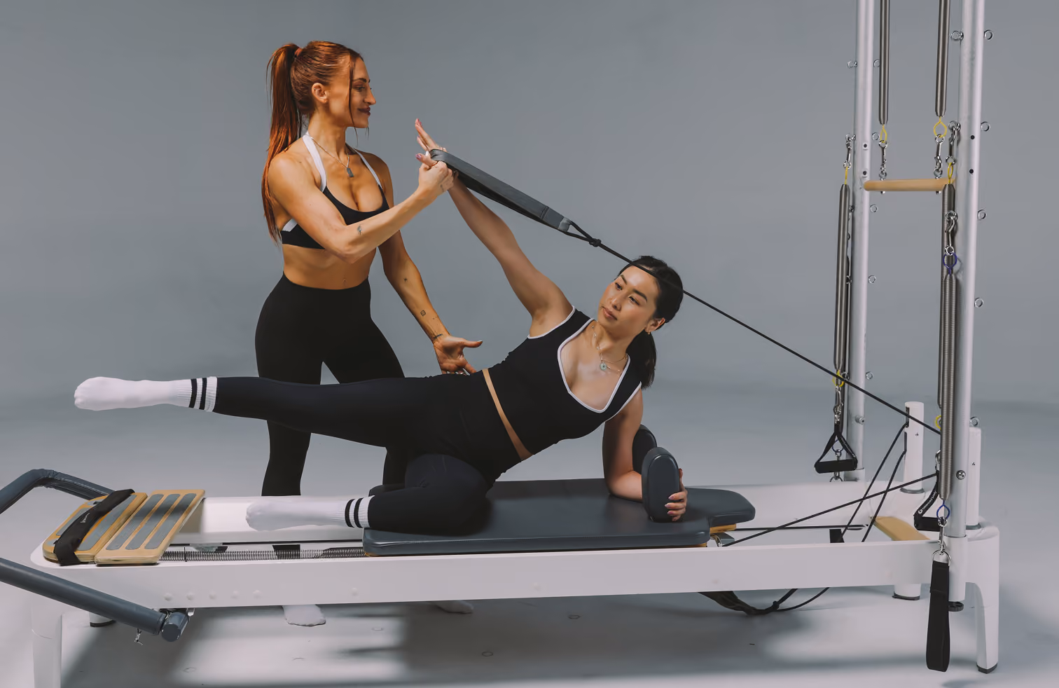 Trainer assists woman doing side leg stretch on Pilates reformer machine in fitness studio.