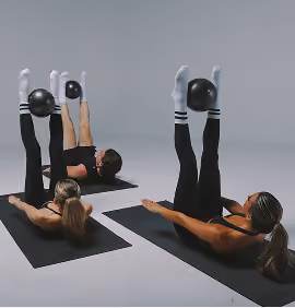 Two women on black mats doing leg raises with black medicine balls held between their feet.