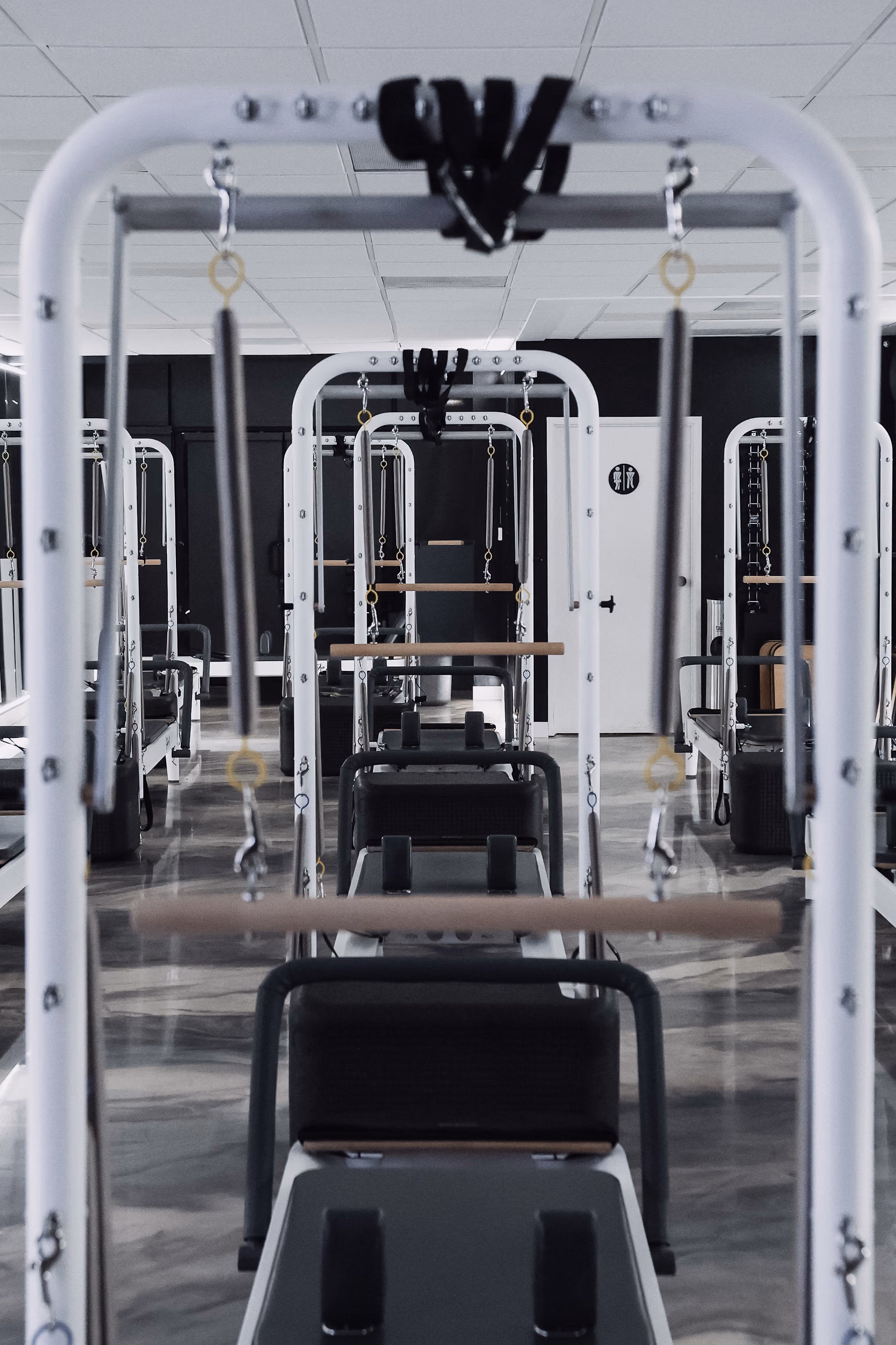 Rows of Pilates reformer machines with white frames and black padded platforms in a clean, modern studio featuring a reflective floor and a white door in the background.