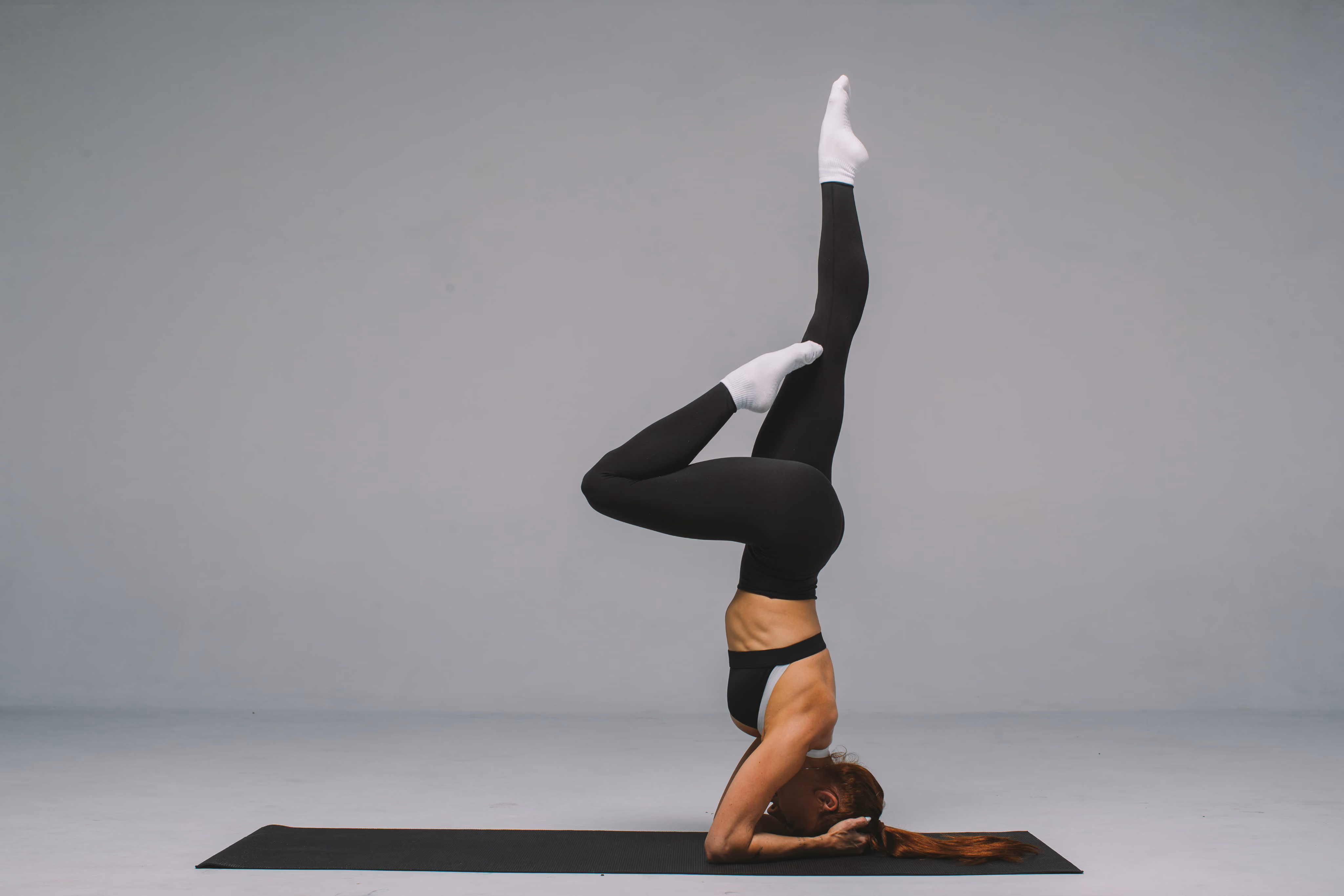 A woman performing a forearm balance yoga pose with one leg bent and the other extended upward, on a black yoga mat against a gray background.