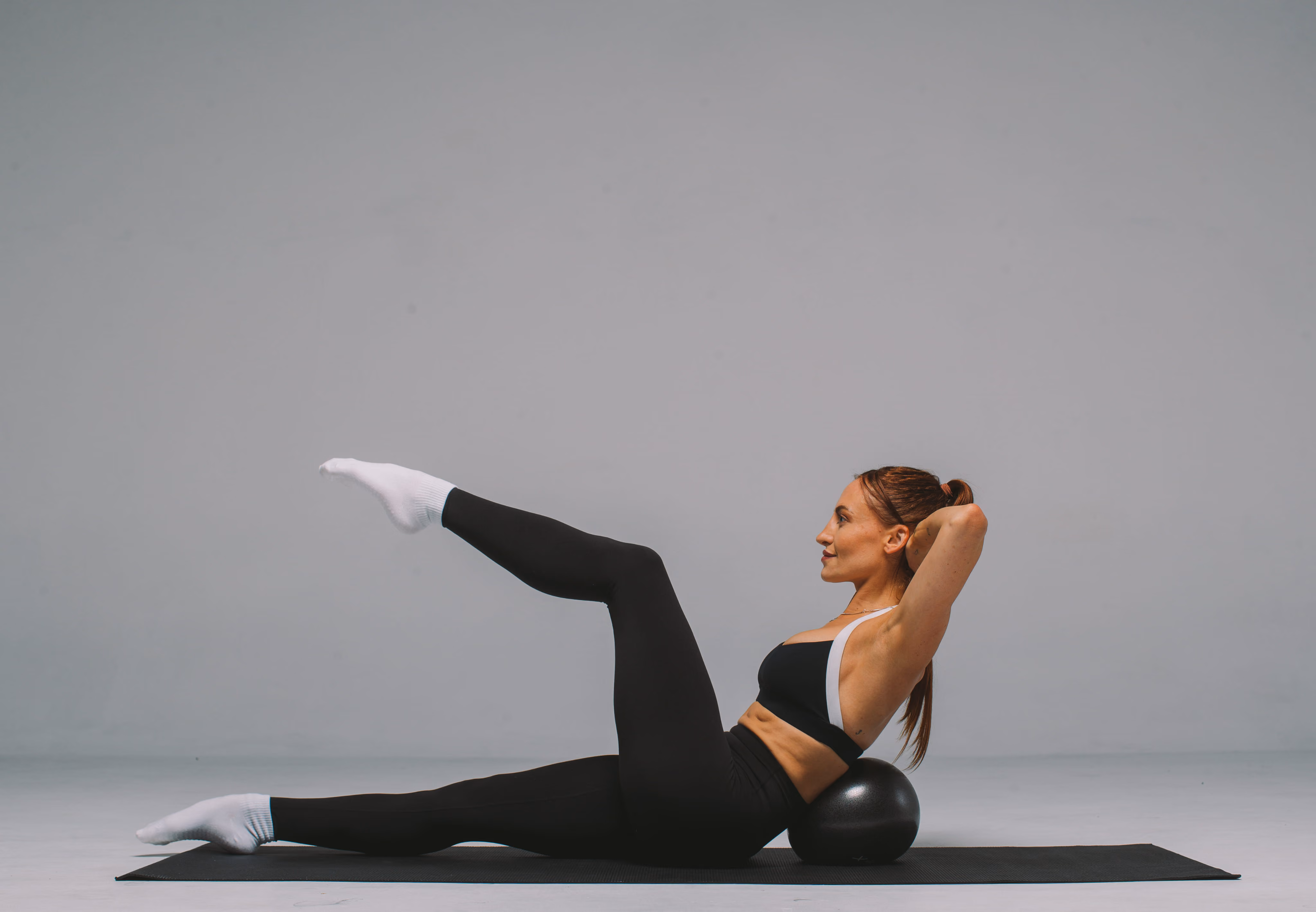 Woman in black workout clothes and white socks exercising on a black mat, balancing her upper back on a black exercise ball with one leg raised and hands behind her head.