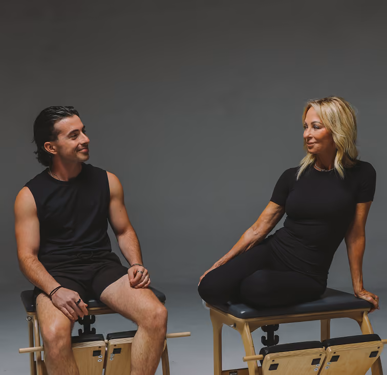A man and a woman dressed in black athletic wear sitting on wooden Pilates chairs, facing each other and smiling against a plain gray background.