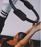 A person with braided hair doing a stretching exercise by holding a black fitness bar overhead while lying down.