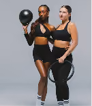 Two women in black athletic wear standing together, each holding a fitness ring, against a plain gray background.