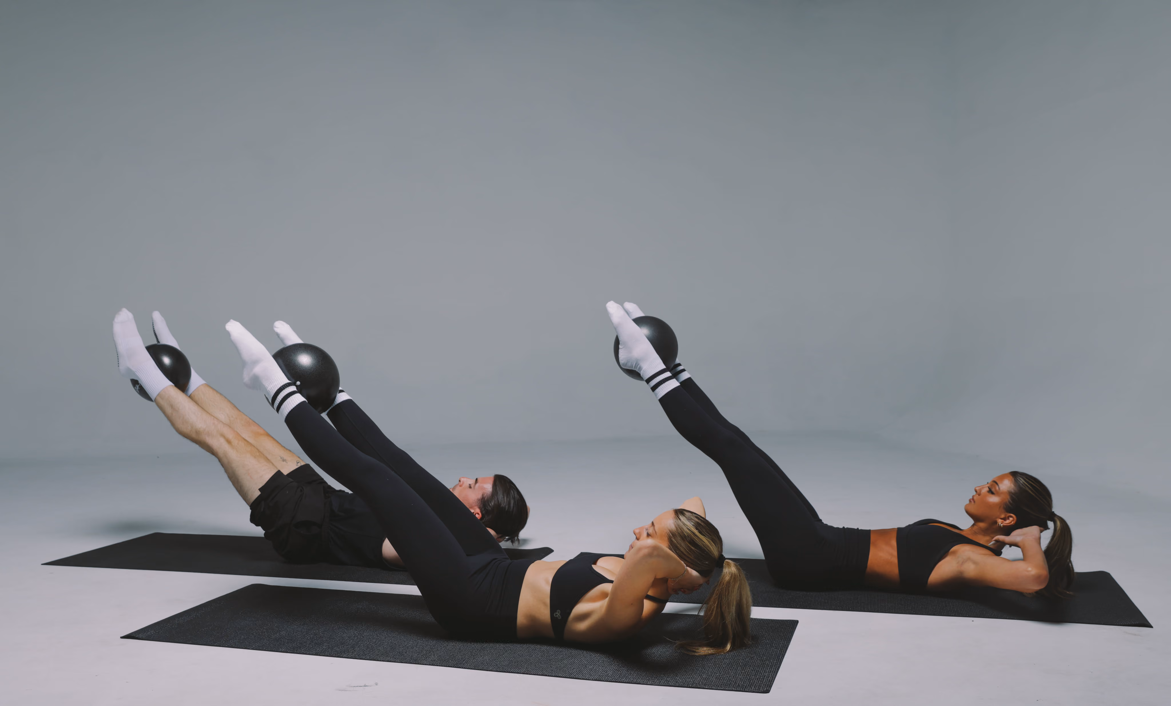Three women lying on exercise mats performing a Pilates or core workout, lifting their legs and holding small black exercise balls between their feet, while supporting their heads with hands.