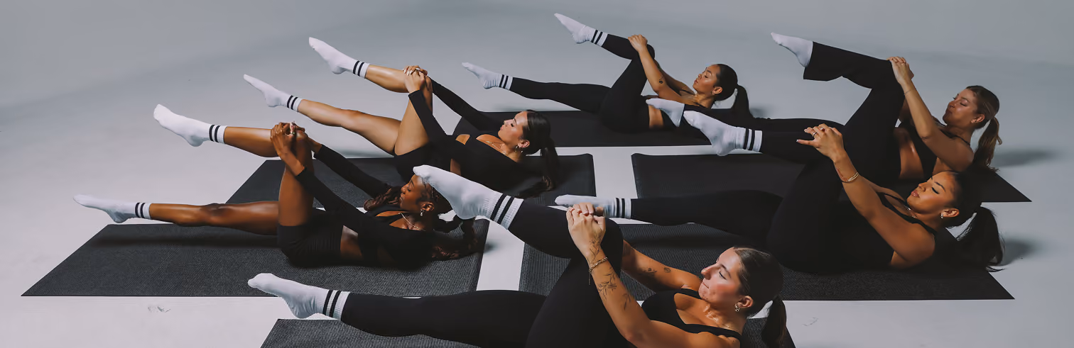 Six women lying on black exercise mats in a studio, wearing black workout clothes and white socks, performing a leg stretch by holding one knee to their chest while the other leg is extended.
