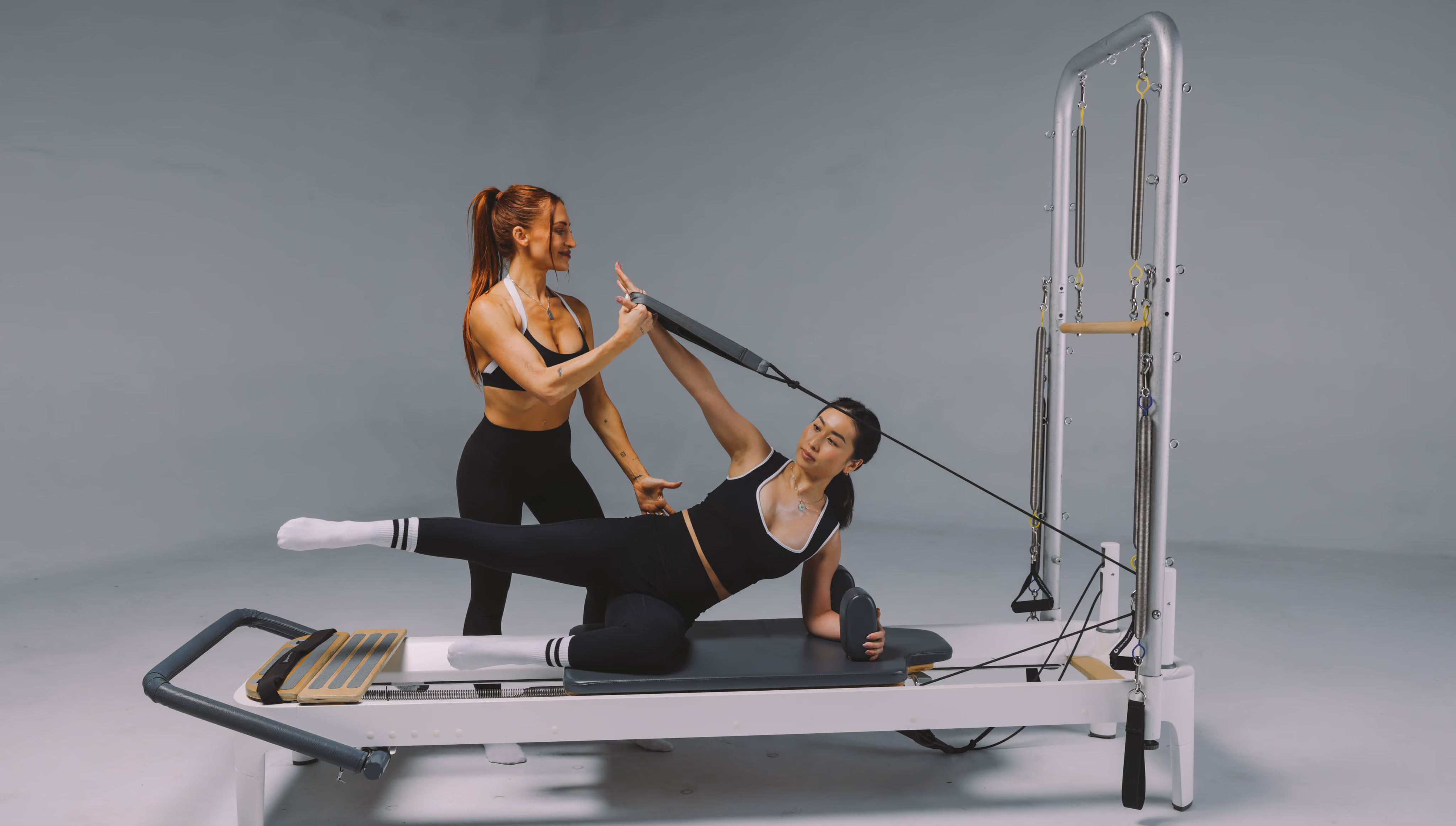 A fitness instructor assists a woman performing a side plank exercise on a Pilates reformer machine, helping her stretch with a strap while she balances on one forearm with one leg extended.