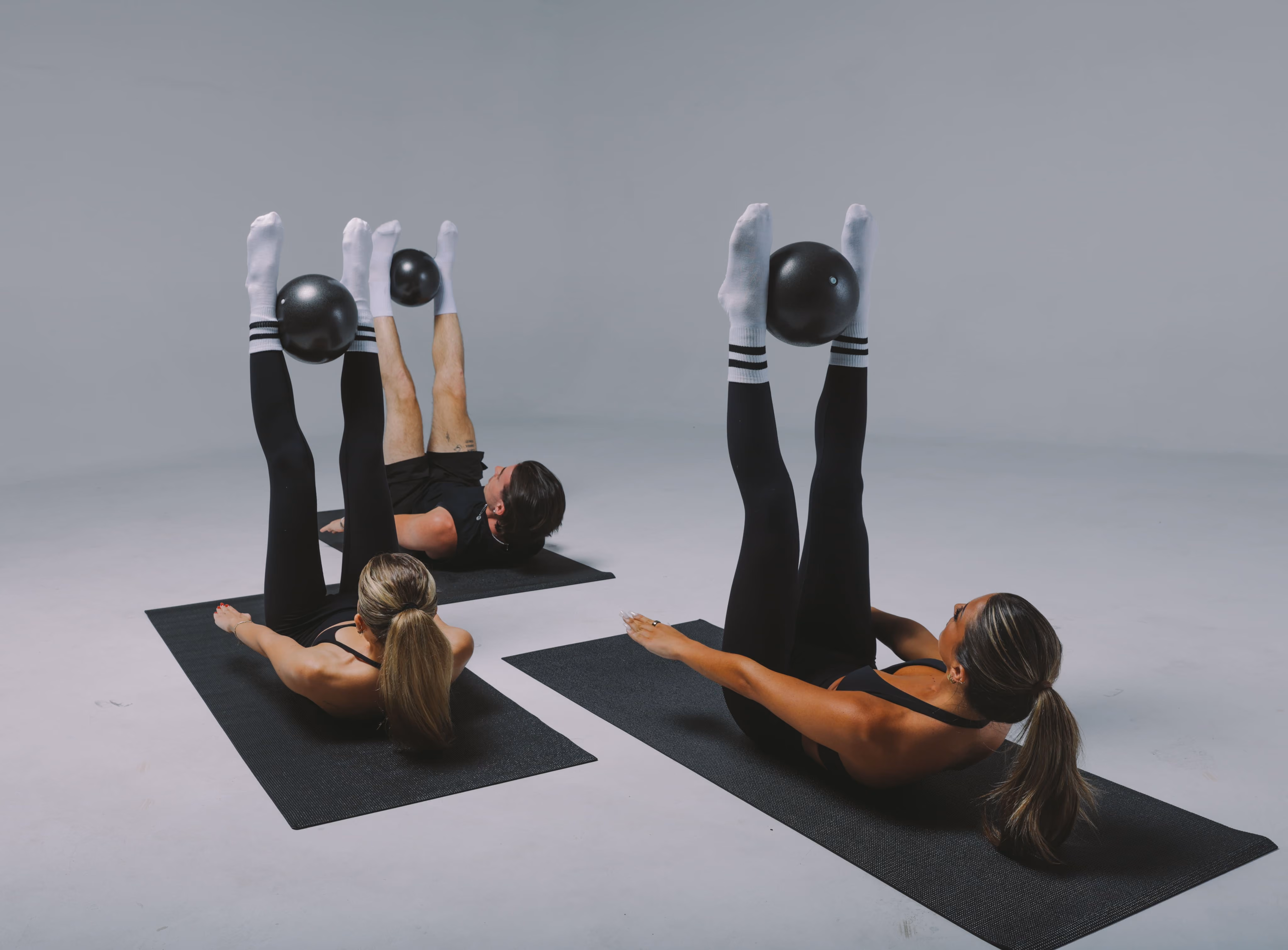 Two women and one man on mats doing leg lifts while holding black exercise balls with their feet.