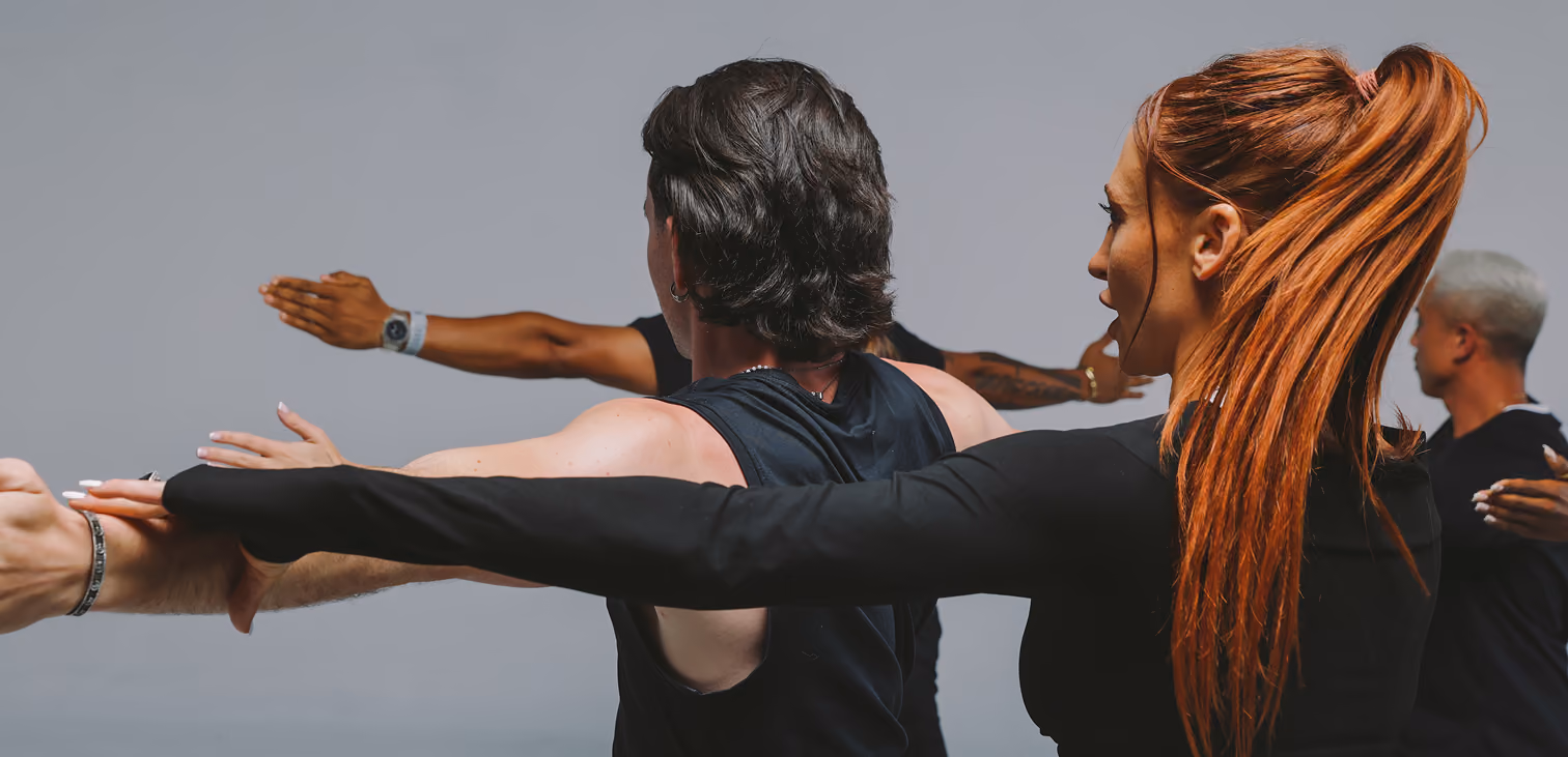 Group of people practicing yoga or stretching exercises with arms extended in a calm indoor setting, focusing on the woman with long red hair in a ponytail.