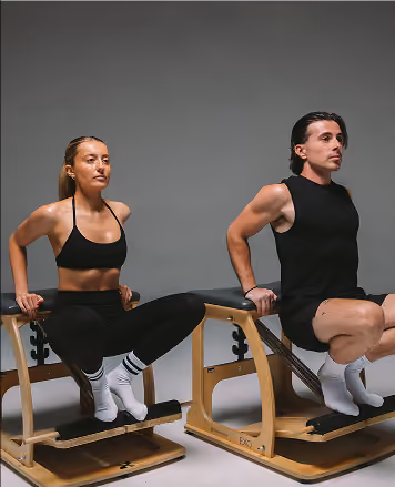 A man and a woman performing tricep dips on wooden exercise benches against a plain gray background, both wearing black athletic clothing and white socks.