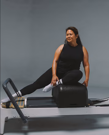 A woman in black workout clothes is stretching on a Pilates reformer machine, with one leg extended and her torso twisted, smiling while exercising indoors.