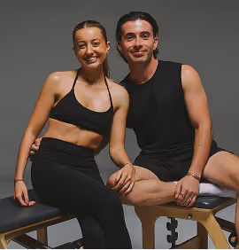 Two women on black mats doing leg raises with black medicine balls held between their feet.