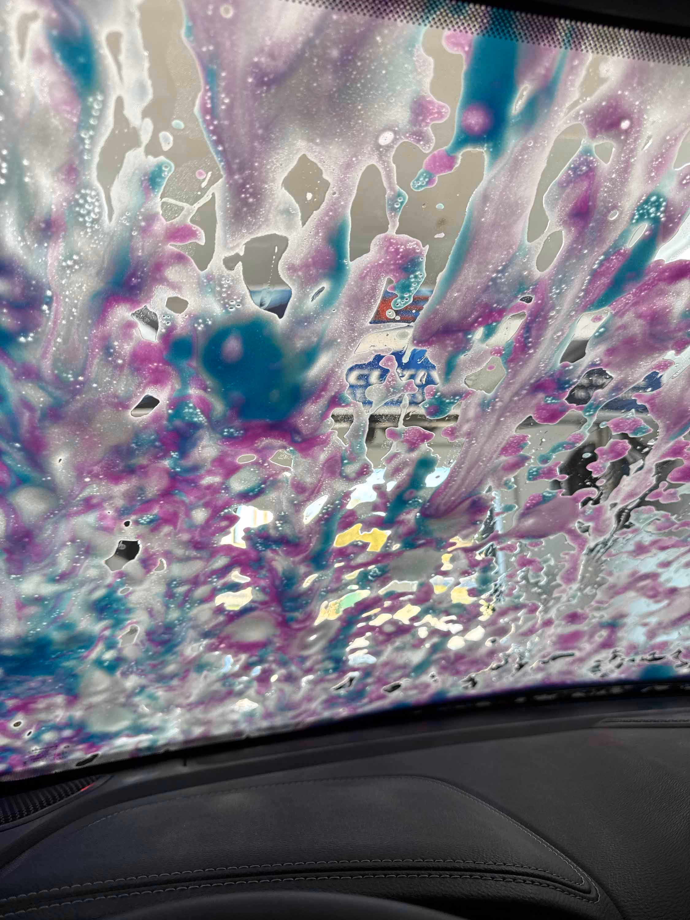 Colorful soap suds covering a car windshield during a wash, visible from inside the car.