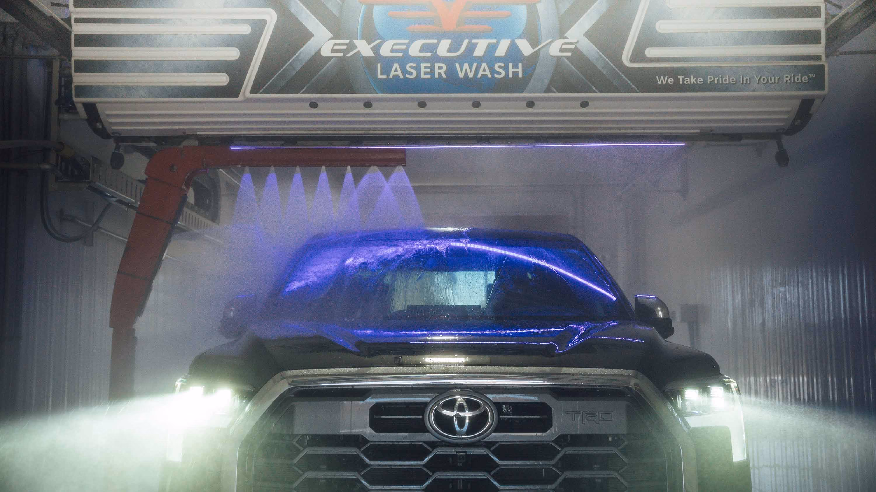 Front view of a black Toyota truck in an Executive Laser Wash with water spraying and headlights on.