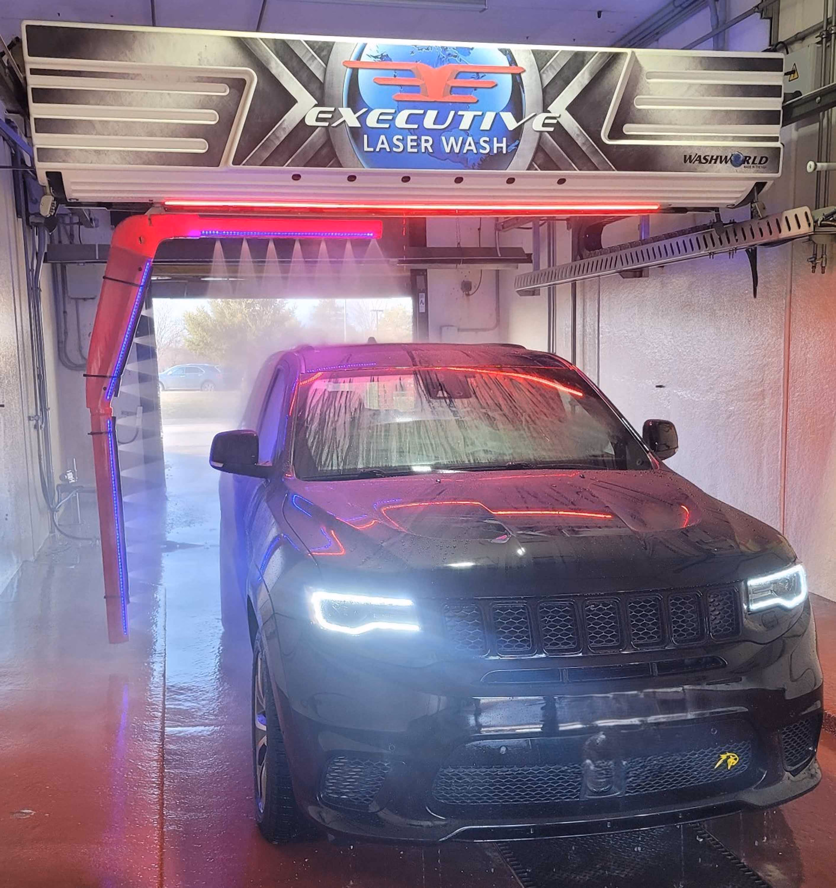 Black SUV with headlights on inside an Executive Laser Wash car wash with purple and red lights.
