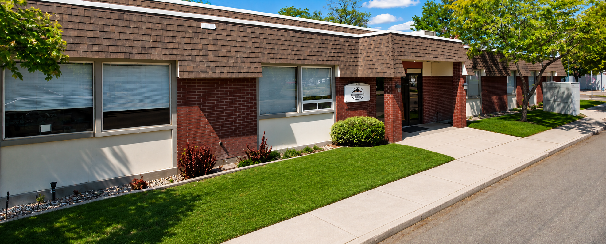 Single-story brick office building with brown shingle roof, large windows with blinds, a glass entrance door, green lawn, small bushes, and trees under a blue sky with scattered clouds.