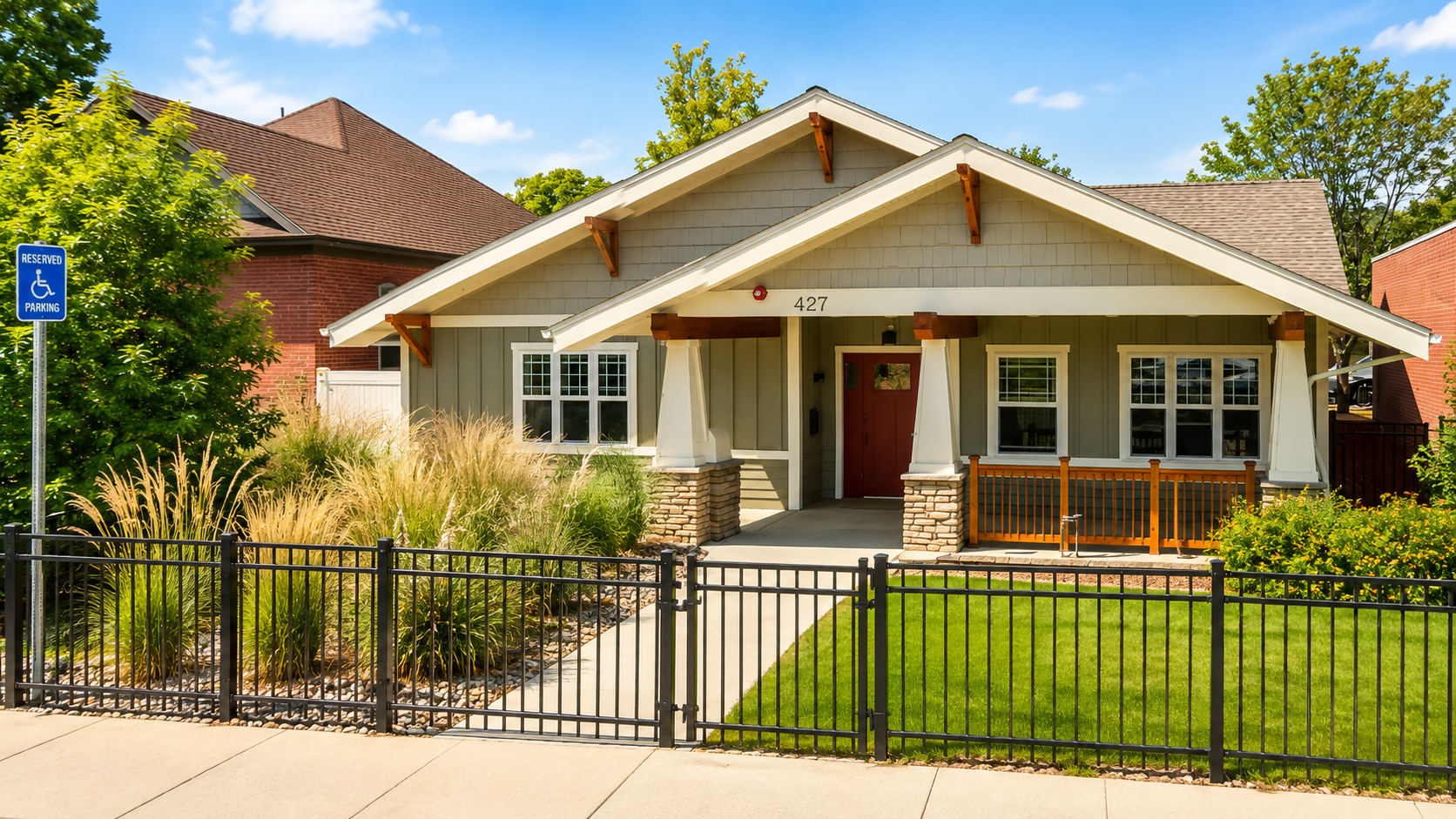 Single-story house with gray siding and a red front door under a gabled roof, featuring stone pillars and a wooden porch railing, surrounded by green grass and landscaped plants behind a black metal fence on a sunny day.