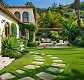 Large garden with stone path, green lawn, lounge chairs, and Mediterranean-style buildings in background.