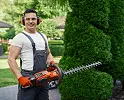 Smiling man holding an orange hedge trimmer standing by a trimmed tall bush outdoors.