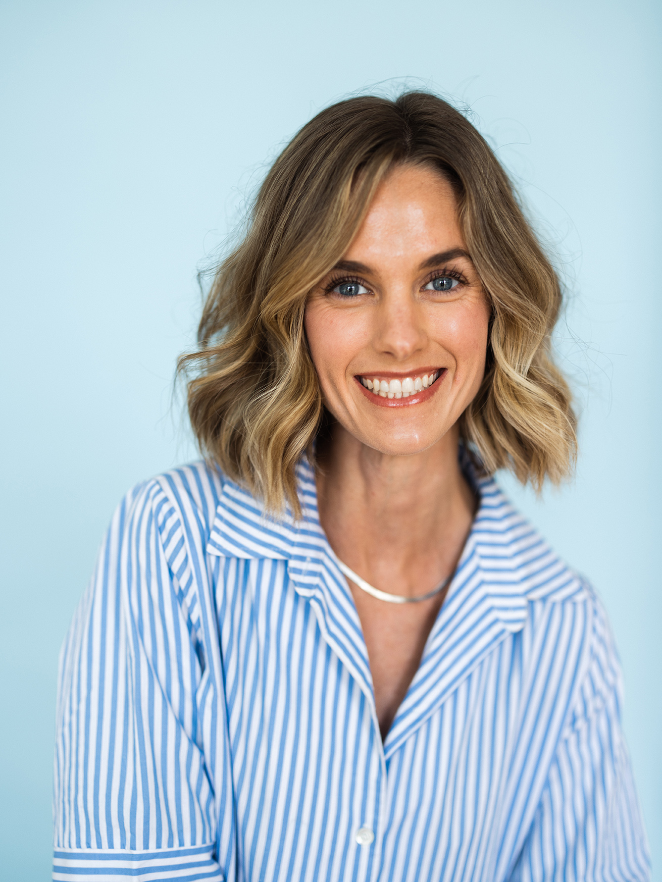 Smiling woman with shoulder-length wavy blonde hair wearing a blue and white striped shirt against a light blue background.