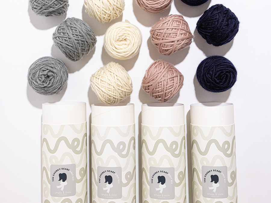 Four cylindrical knitting kit containers labeled 'The Chunky Scarf Knit Kit' with balls of yarn in gray, cream, pink, and navy colors arranged above them on a white background.