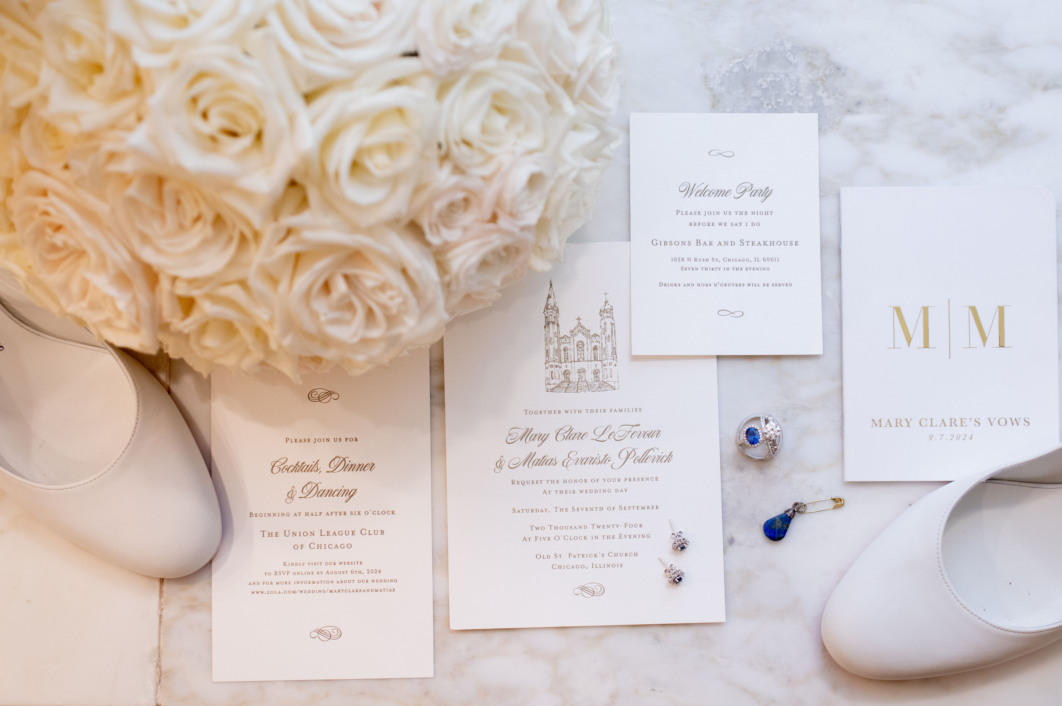 Flat lay of wedding items including white bridal shoes, a bouquet of white roses, wedding invitations, vow booklet dated 9.7.2024, a blue gemstone pendant, diamond earrings, and a sapphire engagement ring on marble surface.