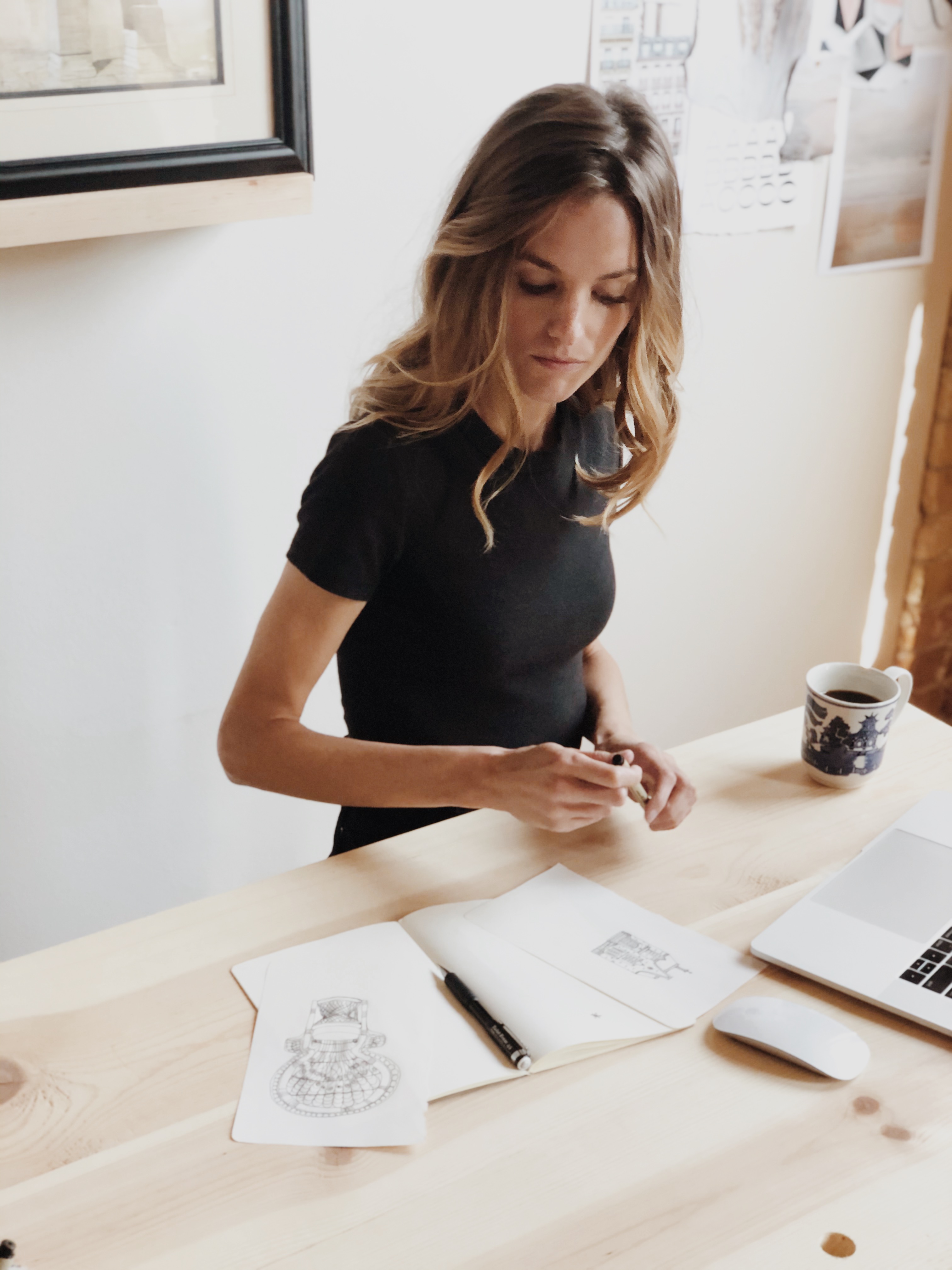 Woman in black shirt sitting at a wooden table, sketching detailed drawings of chairs on paper, with a pen, laptop, wireless mouse, and coffee cup nearby.