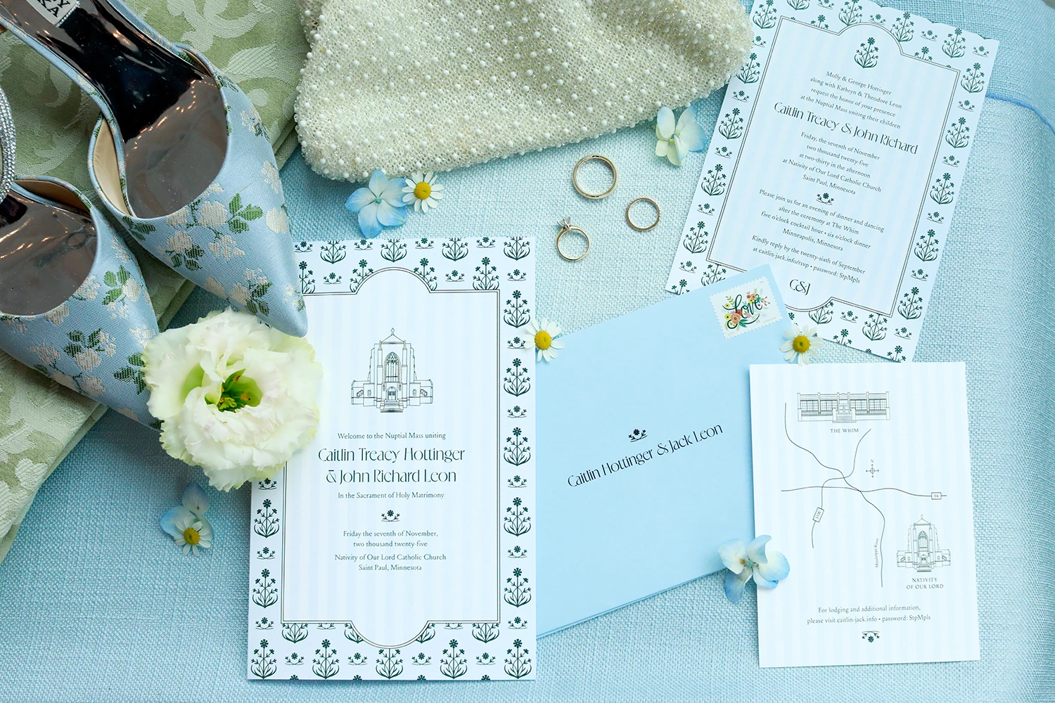 Wedding invitation suite on blue fabric with floral patterned shoes, pearl beaded clutch, white flower, three gold rings, and small blue flowers.