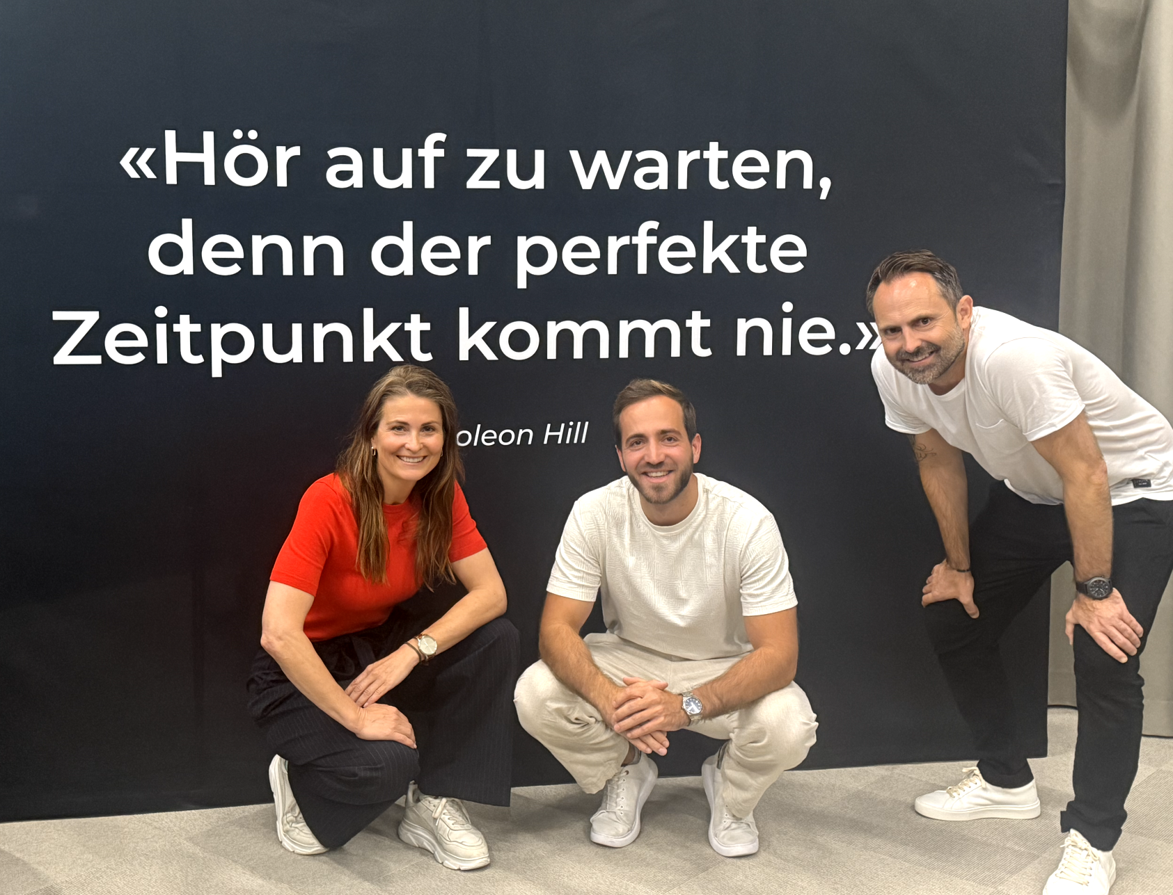 Lukas Hug kniet zwischen zwei Personen vor einer Wand mit dem Zitat Hör auf zu warten, denn der perfekte Zeitpunkt kommt nie von Napoleon Hill