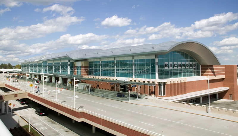 Exterior view of Richmond airport terminal with large glass windows under a curved metal roof and a multi-lane roadway in front under a partly cloudy sky.