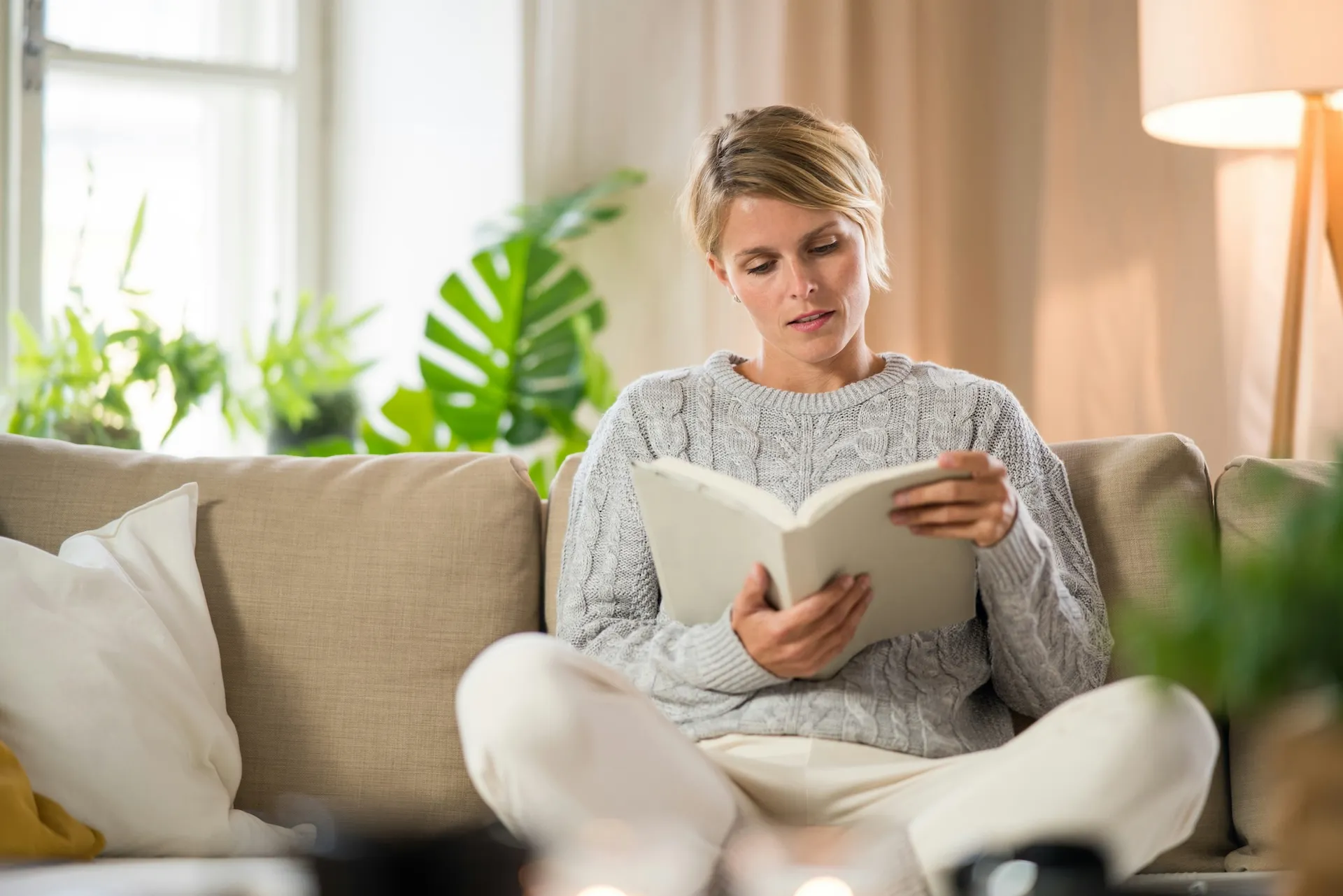 Woman in a gray sweater sitting cross-legged on a couch reading a book in a cozy living room.