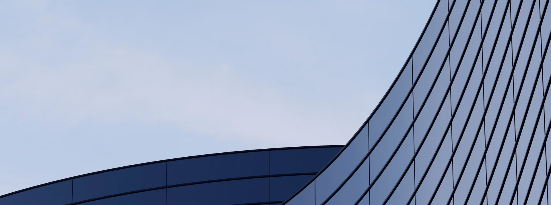 Curved modern glass building facade with reflective windows against a clear blue sky.