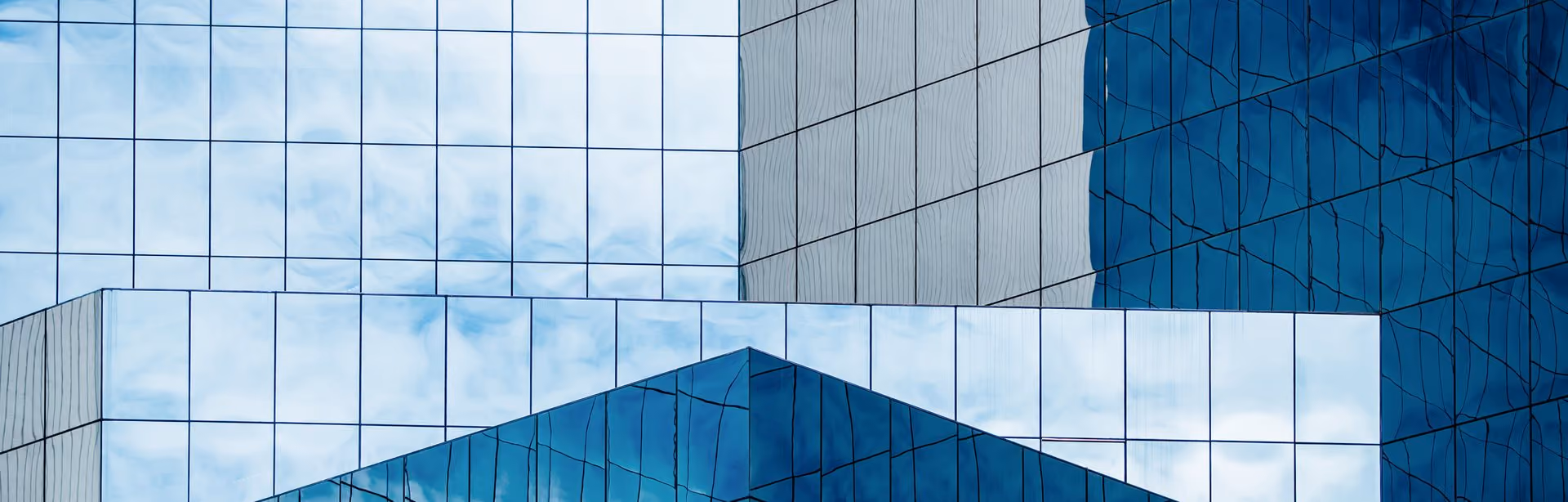 Modern glass building facade with geometric patterns formed by reflective blue-tinted windows and sharp architectural lines.