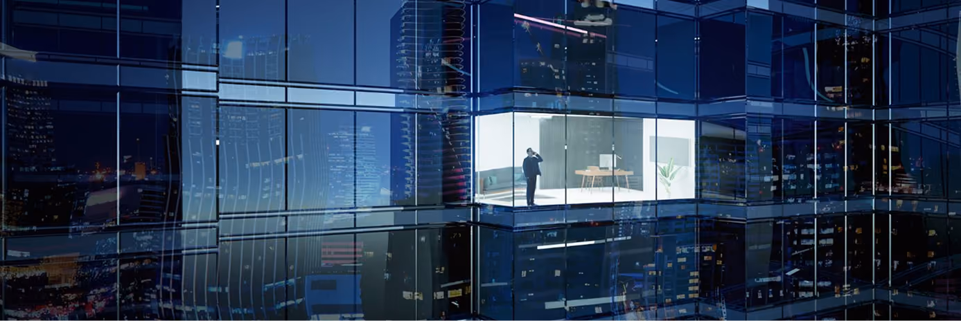 Businessman standing in a brightly lit, modern office with glass walls, seen from outside a dark city building at night with reflections of surrounding skyscrapers.
