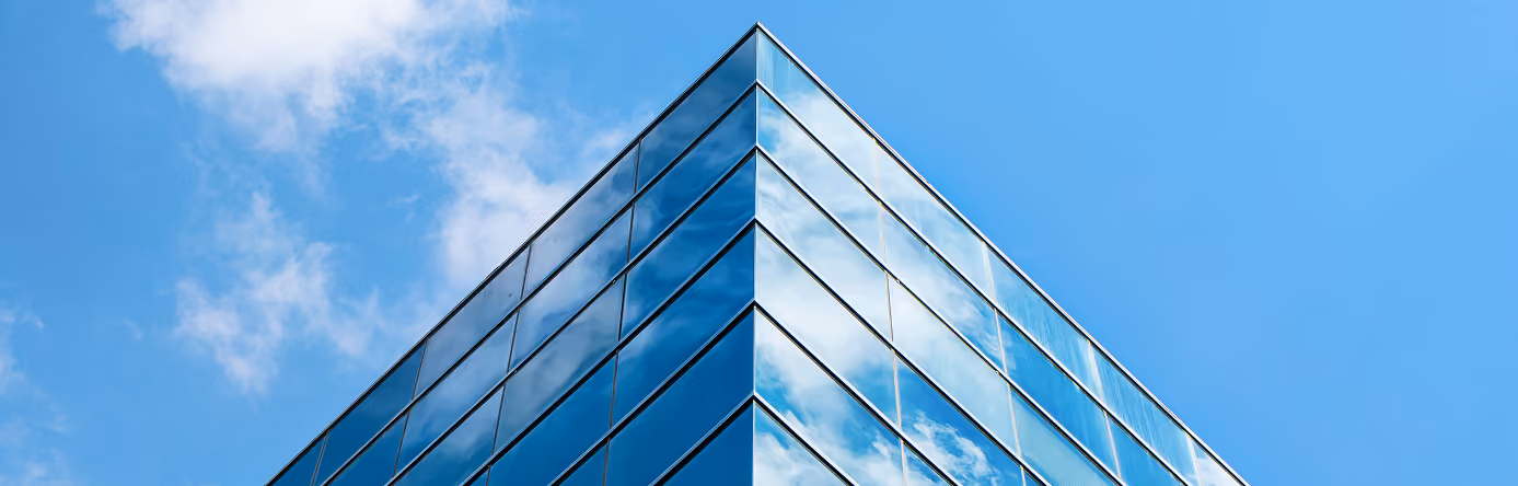 Sharp corner of a modern glass building reflecting clouds against a bright blue sky.