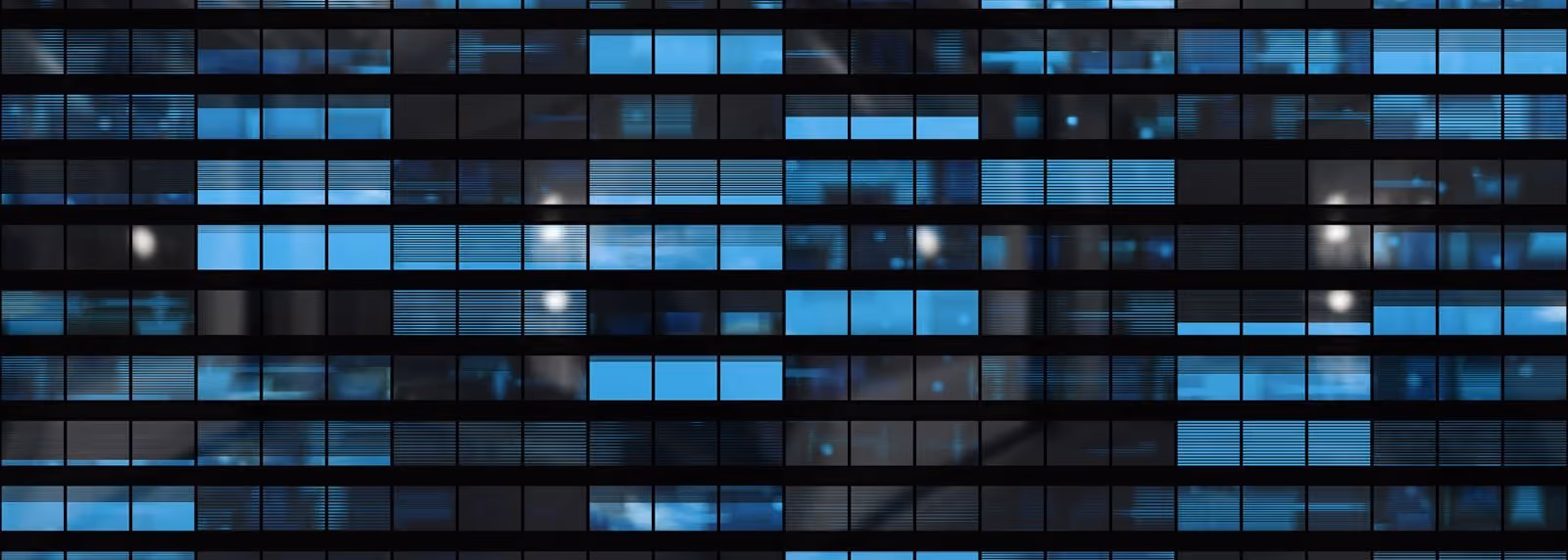 Close-up view of a modern building facade with rows of rectangular windows, some illuminated with blue light and thin horizontal blinds partially closed, creating a grid pattern against the dark structure.