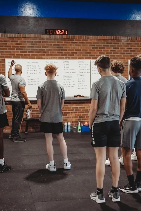 A coach writing on a whiteboard while a group of teen athletes in athletic wear stands and listens during an After School Athlete Development (ASAD) session at Lumen Athletics in Cheyenne, WY.